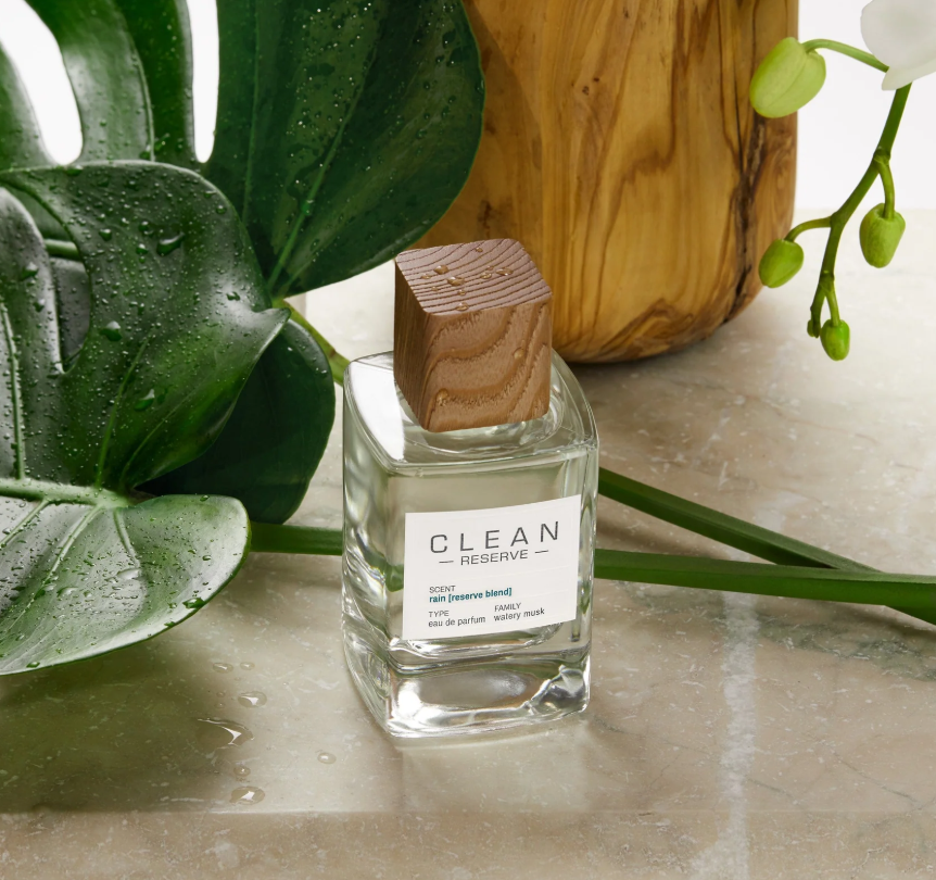 Clean Reserve perfume bottle on a marble surface with green leaves and a wooden block in the background
