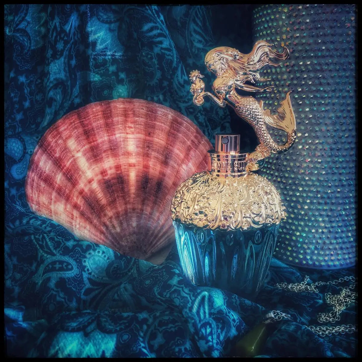 Decorative perfume bottle with mermaid design next to a large shell on a textured blue background